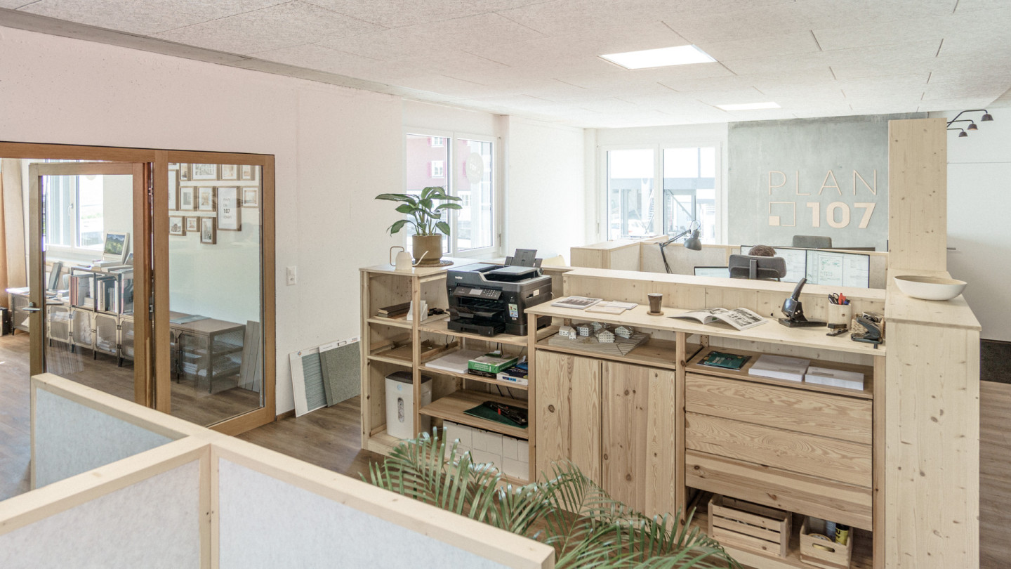 Bürolandschaft der plan107 ag mit Holzmöblierung, Architekturmodellen auf der Theke und Firmenlogo an der Betonwand